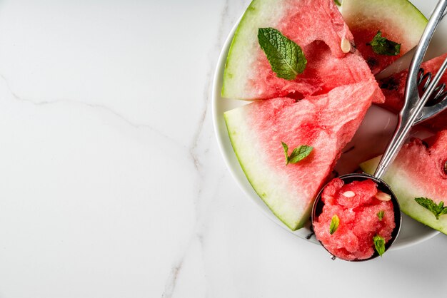 Summer fruit desserts, frozen cocktails. ice cream granite from\
watermelon with mint, one scoop in spoon for ice cream, on plate\
with slices of watermelon. on white marble table. copyspace top\
view