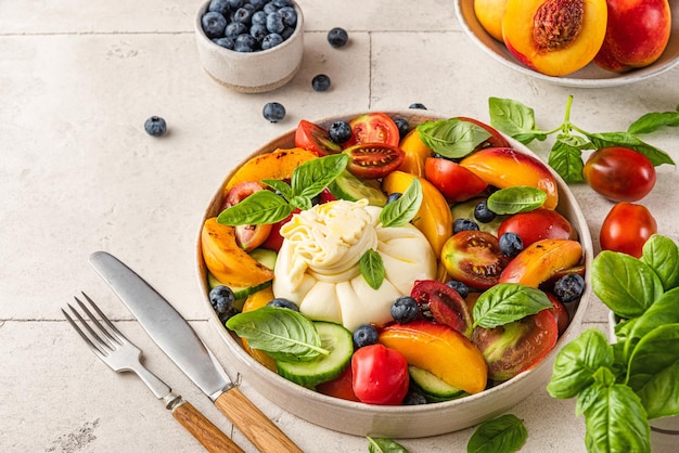 Летний фруктово-овощной салат с сыром буррата и базиликом в тарелке со столовыми приборами на вкусный обед или ужин