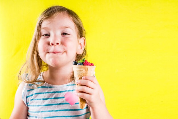 Concetto di vacanza di divertimento di estate, ragazza bionda sveglia del bambino che mangia le bacche dal cono gelato della cialda, fondo giallo luminoso