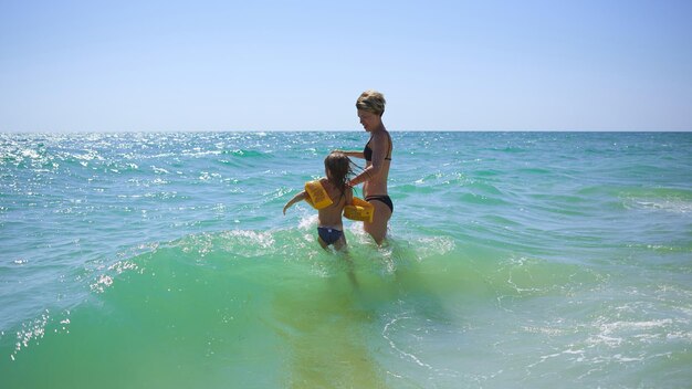 Summer happy family of six years blonde child playing and jumping water waves embracing woman mother in sea shore beach