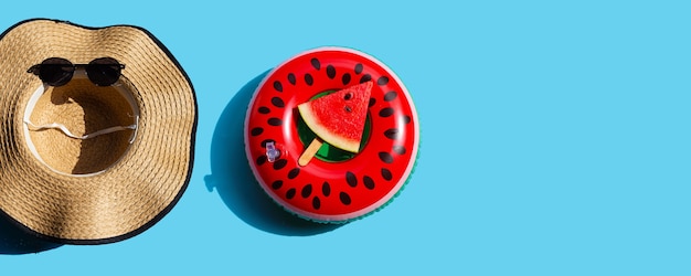 Summer hat and sunglasses with watermelon slice popsicle on swim ring toy on blue background. Summer background concept