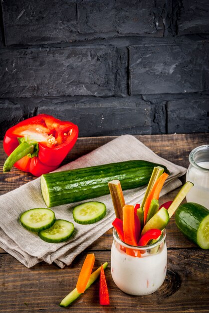 Summer healthy snack appetizer, assorted colorful fresh vegetable sticks (celery, rhubarb, pepper, cucumber and carrot) with yoghurt sauce dip, wood and dark concrete background copy space