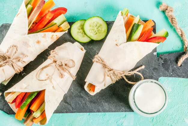 Summer healthy snack, Mexican style tortilla sandwich wraps assorted colorful fresh vegetable sticks (celery, rhubarb, pepper, cucumber and carrot) with yoghurt sauce dip light blue background