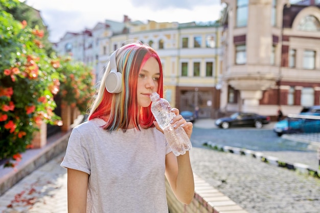 夏、街の暑さ、ボトル入り飲料水を持つ若い女性、街の通りで晴れた日に水を飲むヘッドフォンでティーンエイジャー