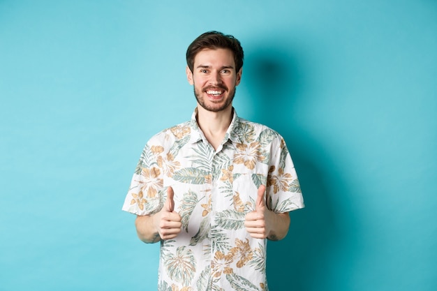 Photo summer holiday. happy tourist showing thumbs up in approval and smiling, wearing hawaiian shirt, recommending travel agency, blue background.