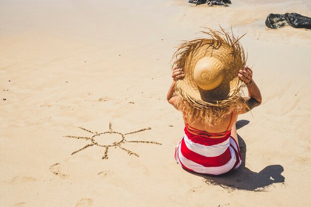Vacanze estive vacanze in spiaggia in paradiso bellissimo resort persone concetto con bella donna con cappello turistico visto dal retro godendosi la sabbia e con il sole disegnato sul suo lato