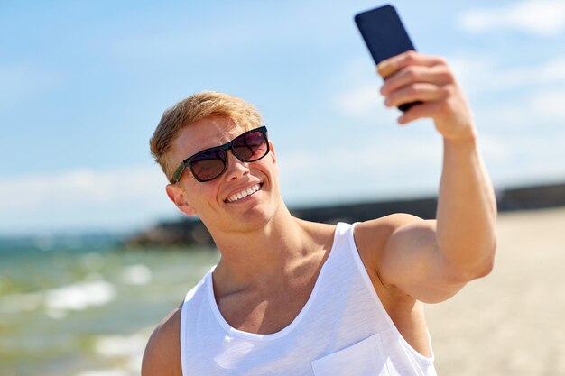 Summer holidays and people concept - happy smiling young man taking selfie by smartphone on beach