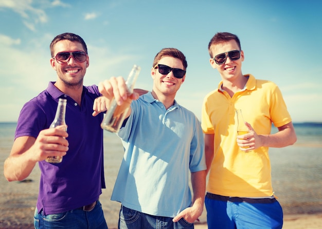 summer, holidays, vacation, happy people concept - group of friends having fun on the beach with bottles of beer or non-alcoholic drinks