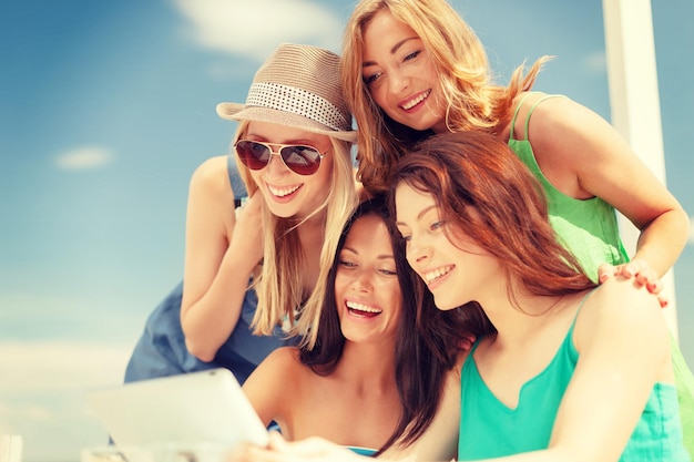 летние каникулы, каникулы, интернет и технологическая концепция - улыбающиеся девушки смотрят на планшетный компьютер в кафе
