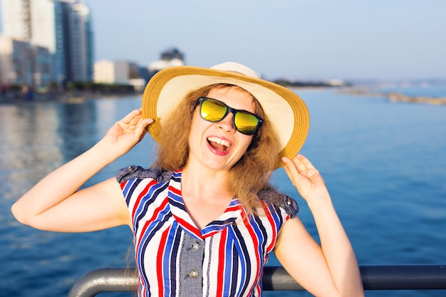 Summer holidays, vacation, travel and people concept - smiling laughing young woman wearing sunglasses and hat on beach over sea background.