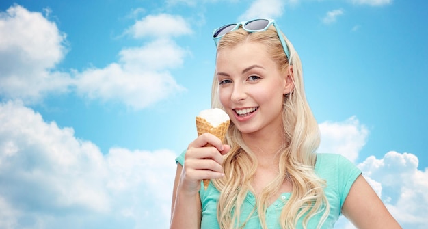 Photo summer, junk food and people concept - young woman or teenage girl in sunglasses eating ice cream over blue sky and clouds background