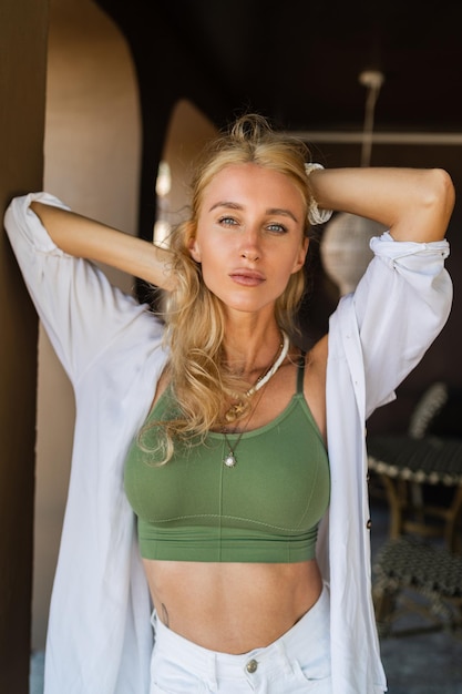 Summer   lifestyle  photo of seductive blond woman with wavy hairs posing  in luxury hotel. Boho asesorises, white blouse and jeans shorts.