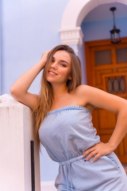 Summer lifestyle portrait of attractive young blonde woman in blue dungarees smiling in a blue house