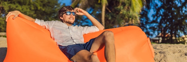 Summer lifestyle portrait of man sitting on the orange inflatable sofa on the beach of tropical