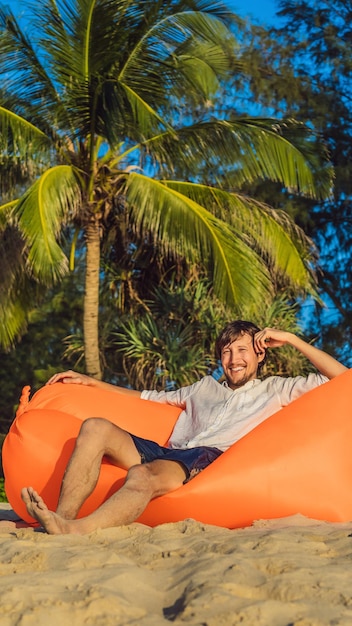 Summer lifestyle portrait of man sitting on the orange inflatable sofa on the beach of tropical