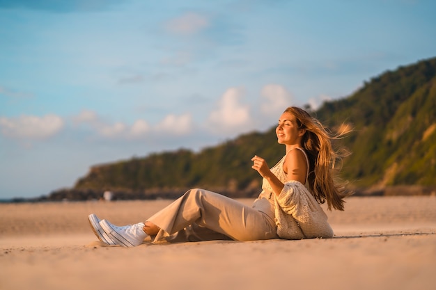 Summer lifestyle, a young blonde with straight hair, in a small wool sweater and corduroy pants sitting on the beach with mountains. Enjoying the soft sea breeze