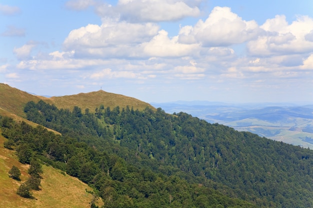 Летний туманный горный пейзаж с зеленым лесом на склоне и христианским крестом на вершине (Украина, Карпаты)