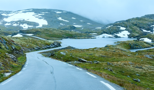 Летний горный туманный пейзаж с дорогой, озером и снегом (Норвегия, Aurlandsfjellet).