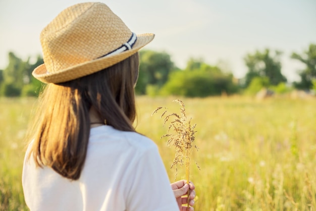 Summer nature, back view of child girl in straw hat tearing meadow grasses, copi space beautiful sunset meadow landscape