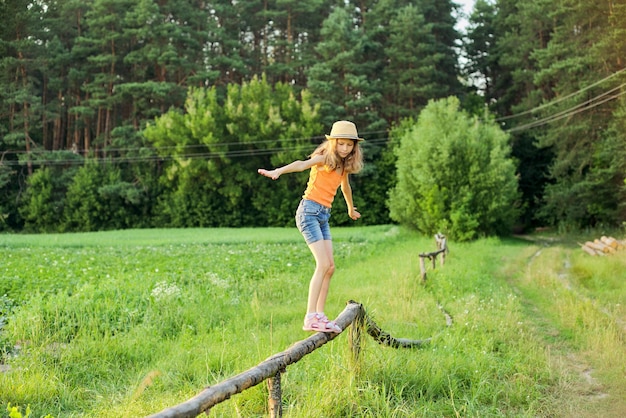 여름, 자연, 행복, 어린 시절 개념. 시골의 나무 울타리를 걷고 있는 모자를 쓴 소녀, 아름다운 일몰 풍경 배경.