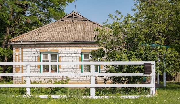 Foto vecchia casa con mattoni a vista estiva nel villaggio. vista di una casa di villaggio tradizionale in campagna dietro un recinto di lavagna bianca e alberi in una giornata di sole estivo.