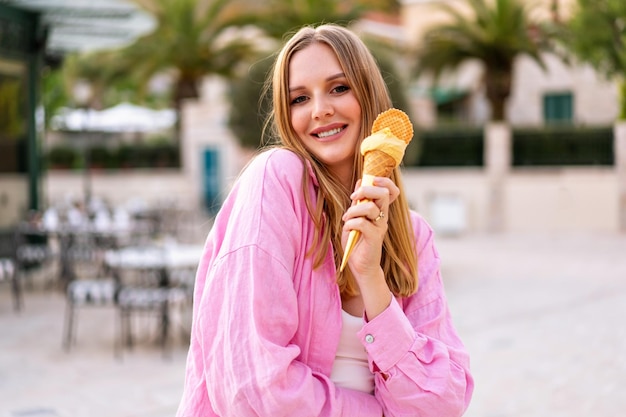 사진 맛있는 이탈리아 젤라토 콘 아이스크림을 먹는 예쁜 금발 여성의 여름 야외 초상화는 휴가 핑크색 트렌디한 의상을 즐긴다