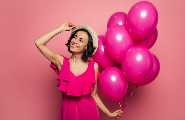 Summer party. Close-up photo of a smiling girl who is touching her straw hat with her right hand and holding pink balloons in her left hand, while looking aside.