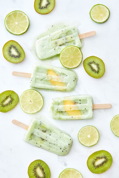 summer pattern of frozen lime kiwi juice on a stick with a piece of peach and pieces of fruit on a gray background. healthy dessert. flat lay