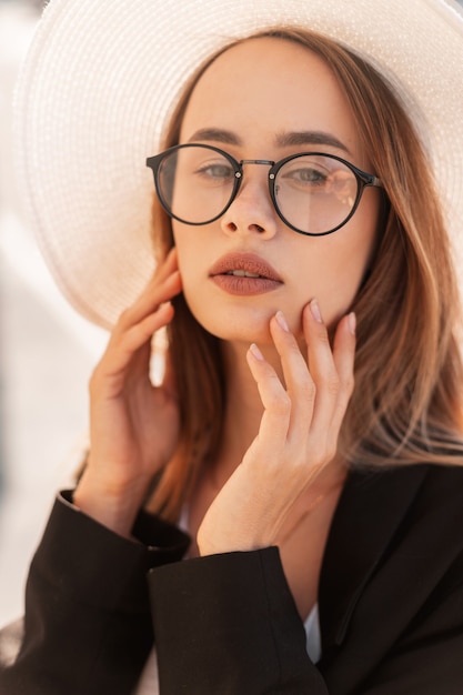 Summer portrait beautiful fashionable young woman in fashion glasses in straw white hat in youth black stylish jacket at sunset. Elegant girl on street in sunny day. Trendy summer clothes for women.