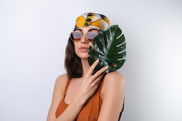 Летний портрет молодой женщины в спортивном купальнике, платке и солнцезащитных очках с пальмовым листом монстера