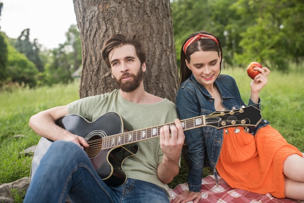 Estate, riposo. giovane ragazzo barbuto che suona la chitarra per una ragazza con una mela seduta su una coperta sotto un albero all'aperto nel bel pomeriggio