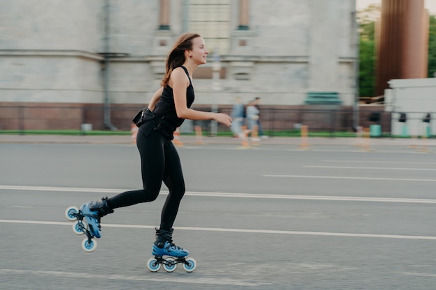 Summer rollerblading and fitness concept. Fit young dark haired woman fond of rollerskating exercises all body poses on road along asphalt during sunny day wears comfortable clothes has sporty figure