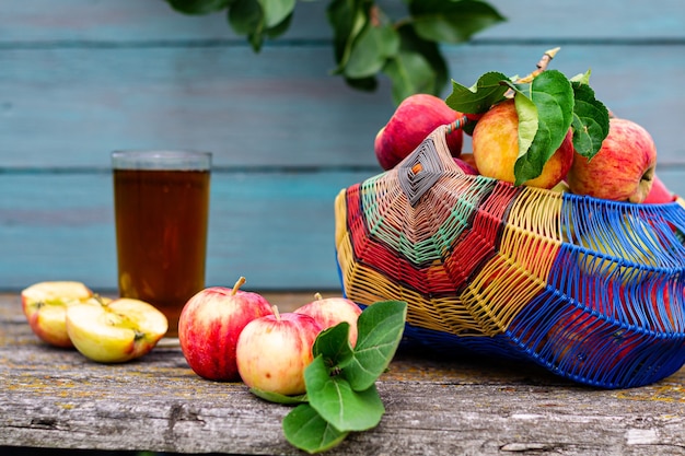 Summer rustic still life, a basket of ripe garden apples on a wooden background, apple juice in a transparent glass. Seasonal harvest concept.