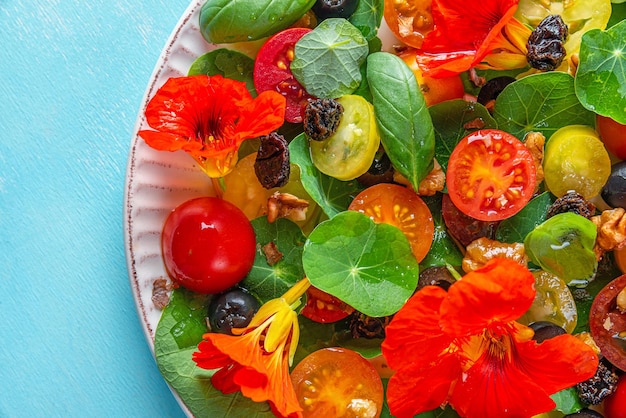 Summer salad with nasturtium flowers tomatoes olives raisins and nuts on blue background Close up Top view