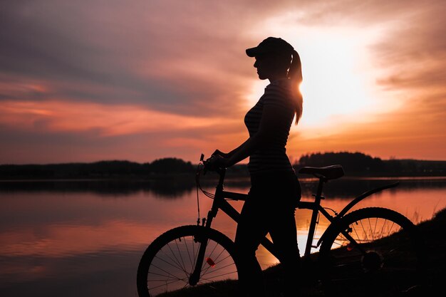 夕日を背景に湖のほとりに自転車を持った夏のシルエット風景少女真っ赤な夕日