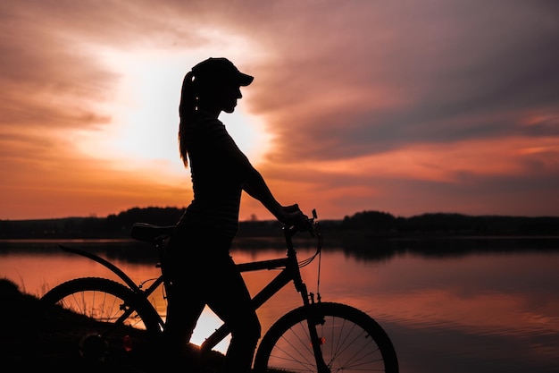 夕日を背景に湖のほとりに自転車を持った夏のシルエット風景少女真っ赤な夕日