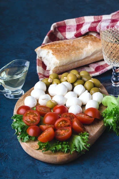 Spuntino estivo di mozzarella, pomodori, olive e verdure su tavola di legno, con bicchiere di vino bianco, pezzo di baguette, olio d'oliva e tovagliolo a scacchi rosso. sfondo blu scuro