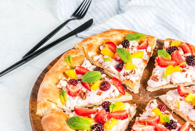 Summer snacks. Food for party. Fruit pizza with cream, currants, yogurt, strawberries, mango, peaches, bananas, blackberries, chocolate, walnuts, mint. On light blue table. copyspace