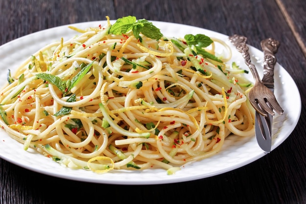 Summer spaghetti alla carrettiera with mint leaves lemon zest pecorino cheese chili peppers flakes parsley and grated zucchini on a white plate on a dark  table with cutlery