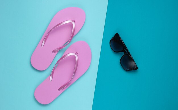 写真 夏の静物。ビーチアクセサリー。ファッショナブルなビーチピンクのビーチサンダル、青い紙の背景にサングラス。フラットレイ。上面図