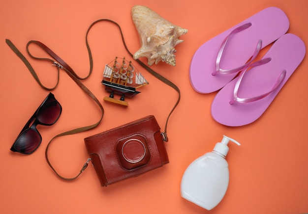 Summer still life. Beach accessories. Fashionable pink flip flops, retro camera, sunblock bottle, sunglasses, seashell on coral paper background.