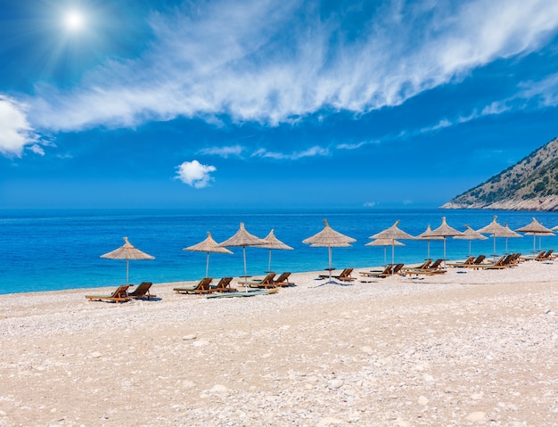 Летний солнечный пляж с аквамариновой водой и облаками в небе, шезлонгами и соломенными зонтами от солнца (Албания).