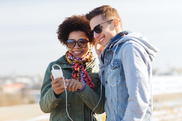 лето, технологии, люди и концепция дружбы - улыбающаяся пара со смартфоном и наушниками, слушающая музыку на городской улице