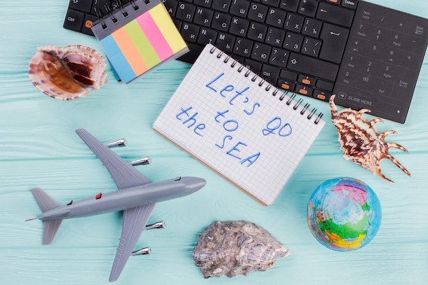 Summer travel accessories consist of seashells, airplane, globe\
on blue background. let\'s go to sea on notebook.