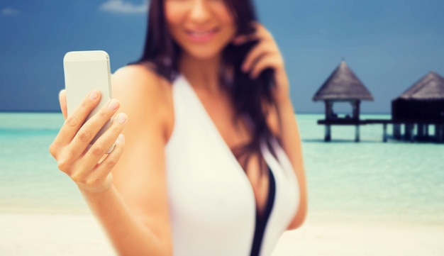 summer, travel, technology and people concept - close up of sexy young woman taking selfie with smartphone over bungalow on beach background