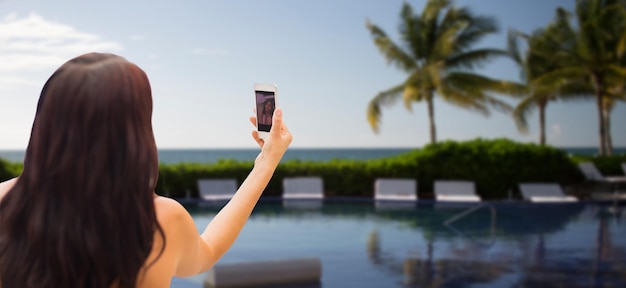 summer, travel, technology and people concept - close up of sexy young woman taking selfie with smartphone over resort beach with palms and swimming pool background