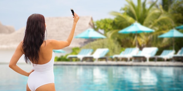 summer, travel, technology and people concept - sexy young woman taking selfie with smartphone over beach and swimming pool background