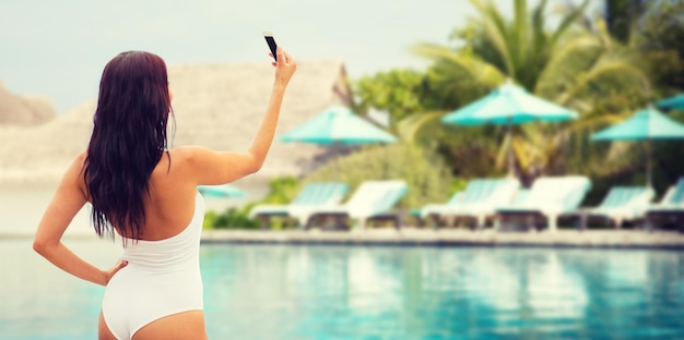 Foto concetto di estate, viaggio, tecnologia e persone - giovane donna sexy che prende selfie con lo smartphone su sfondo spiaggia e piscina