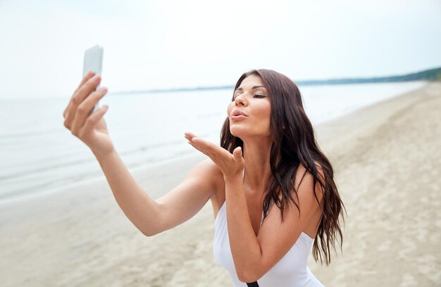 summer, travel, technology and people concept - sexy young woman taking selfie with smartphone and sending blow kiss on beach