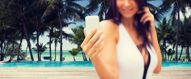 лето, путешествия, туризм, технологии и концепция людей - крупный план сексуальной молодой женщины, делающей селфи со смартфоном над пляжем курорта с пальмами и фоном бассейна